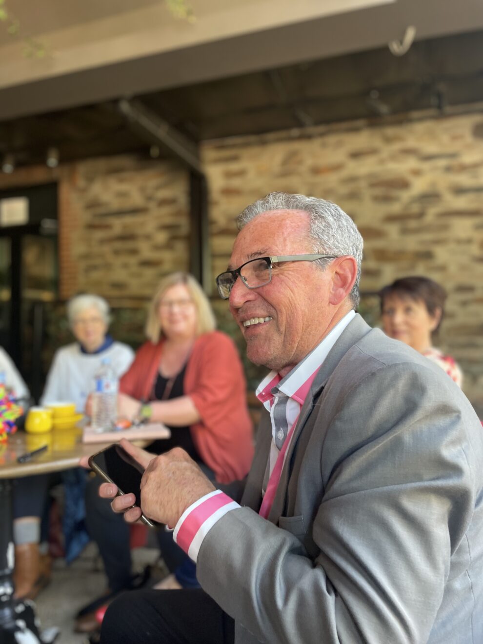 A man with great hair and wearing a grey jacket and red and white striped shirt smiles to someone off camera. A group of people are visible at cafe tables in the background.