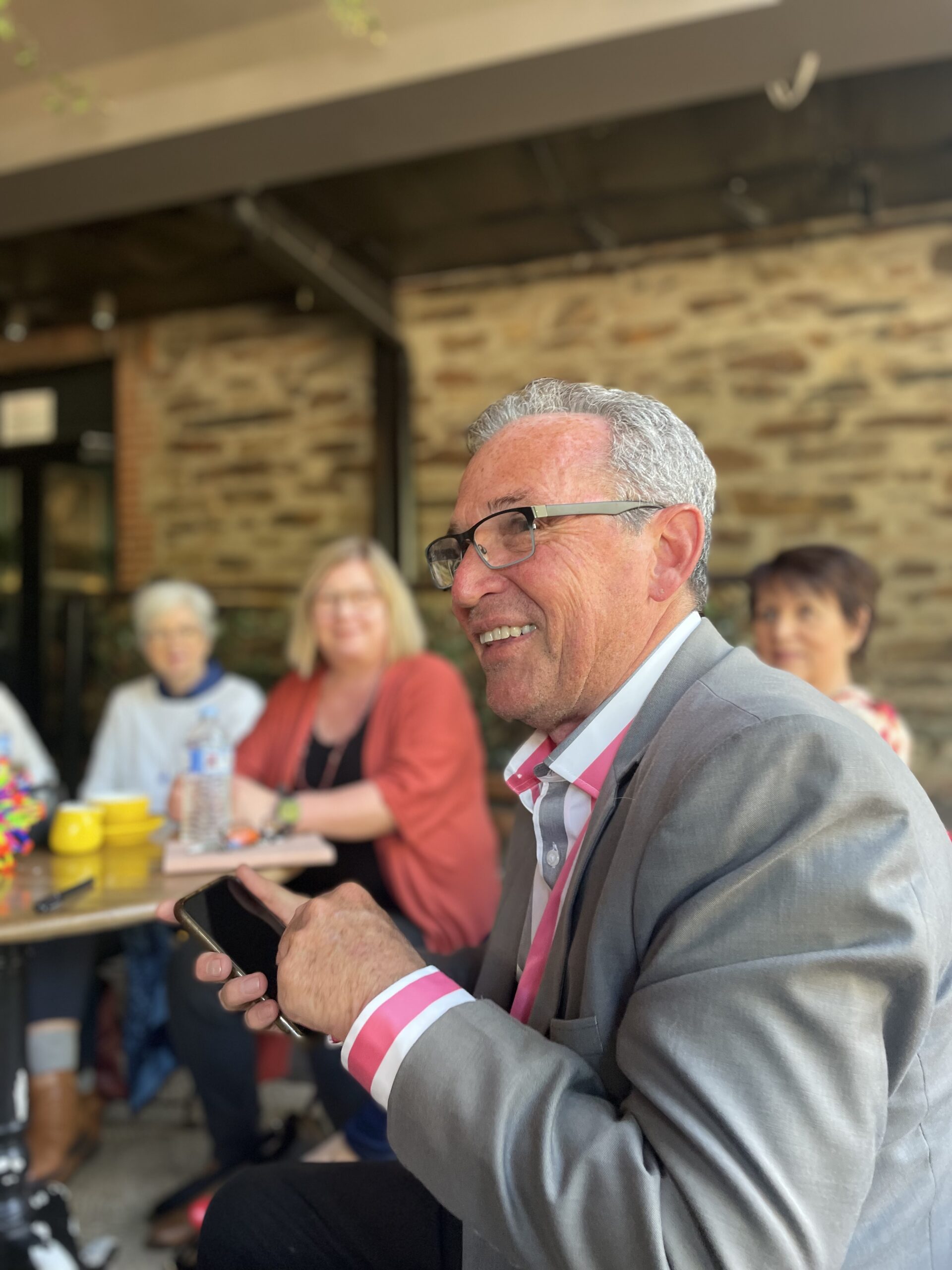 A man with great hair and wearing a grey jacket and red and white striped shirt smiles to someone off camera. A group of people are visible at cafe tables in the background.