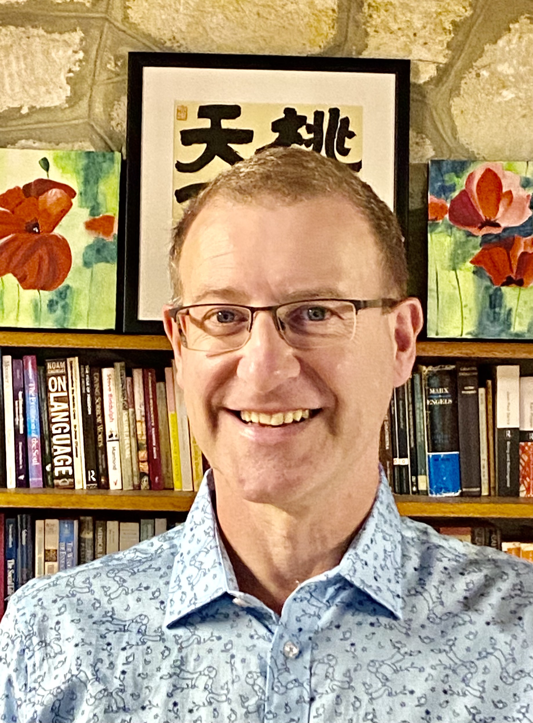 A man wearing glasses and wearing a blue floral shirt stands smiling at the camera in front of a bookshelf.