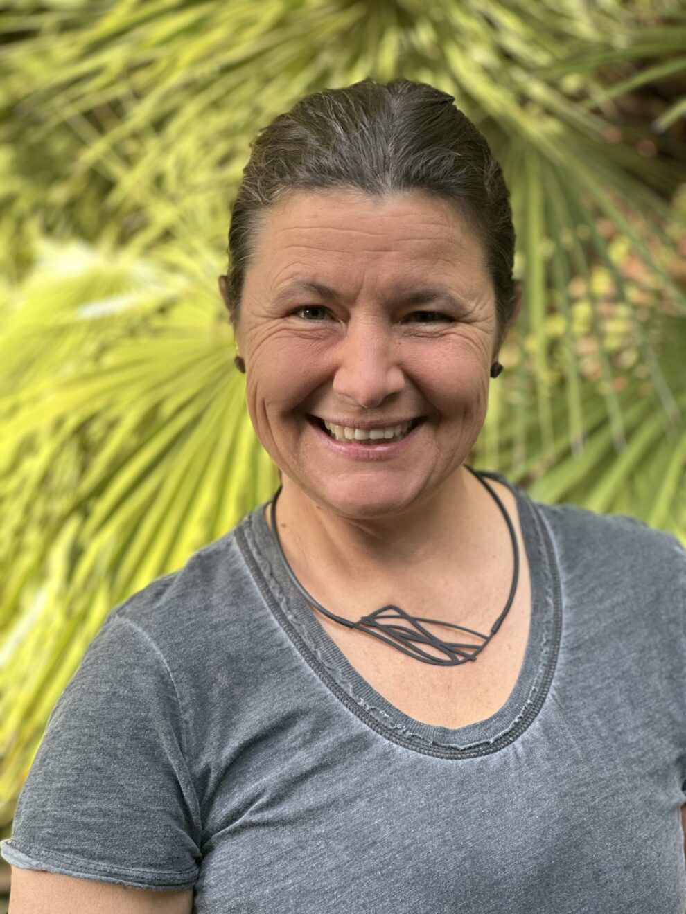 a woman with dark hair tied back in a pony tail and wearing a grey top in front of bright green palm leaves.