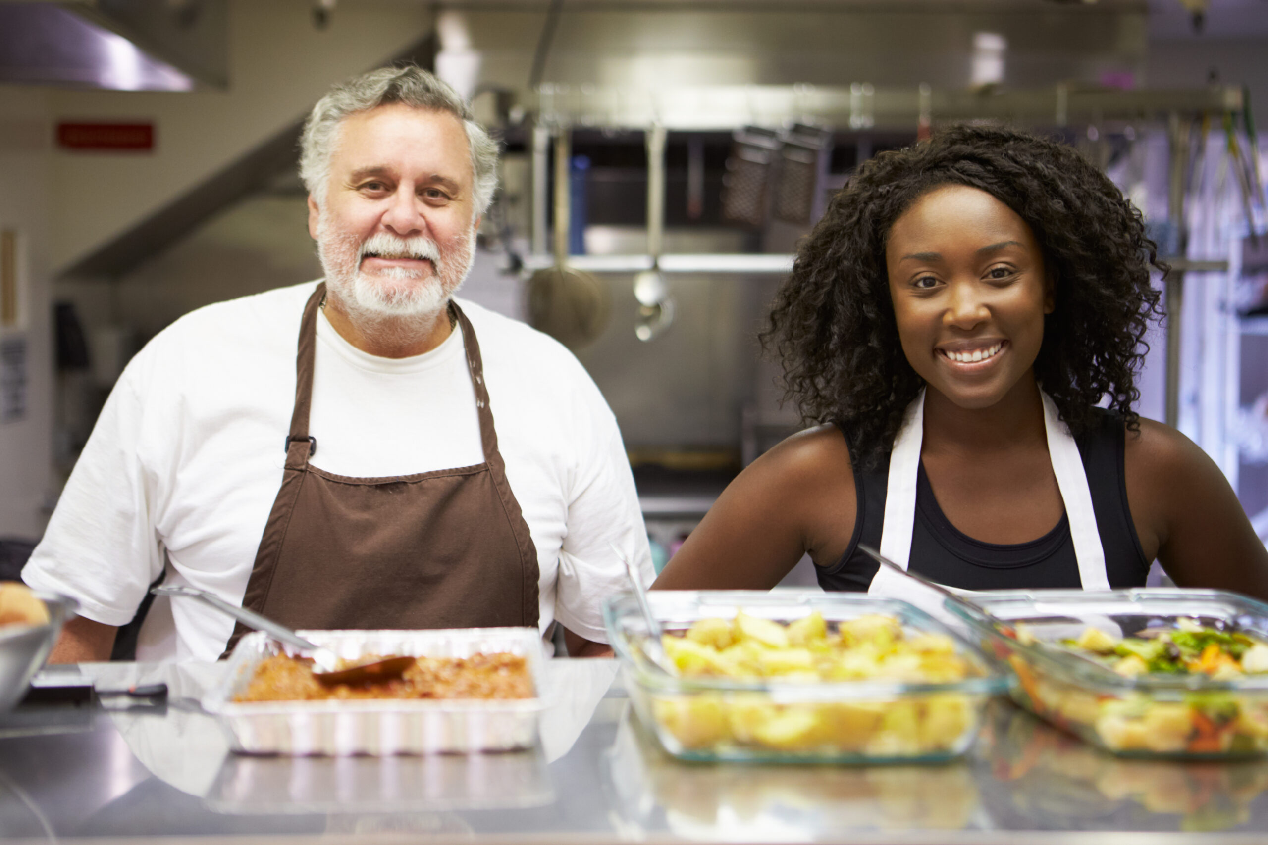 An older man and a younger woman from different racial backgrounds stand side by side in a commercial kitchen smiling at the camera.