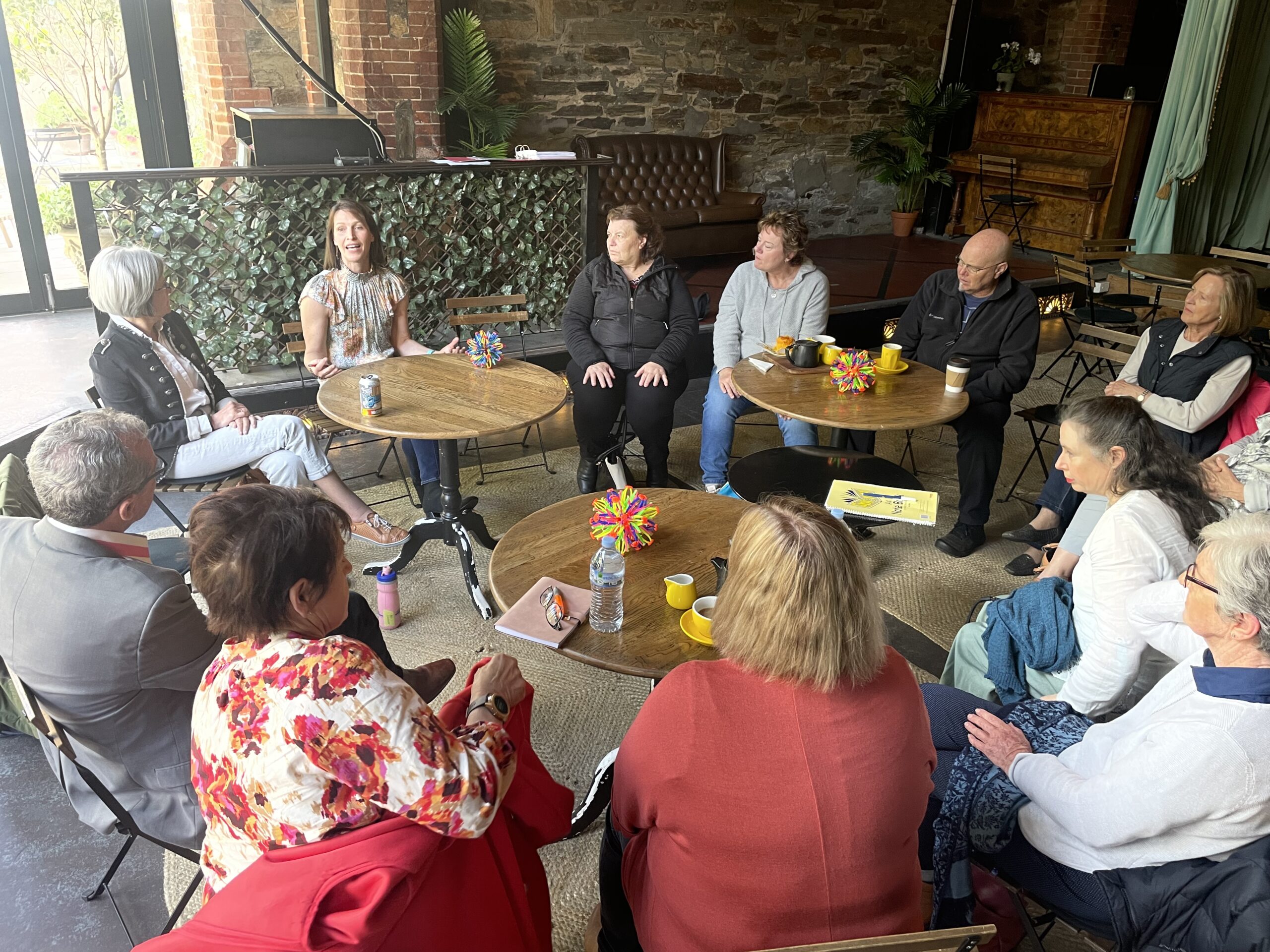 a group of people sit in a circle around cafe tables, listening to a woman who is talking to the group