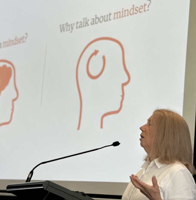 A head and shoulder side view of a woman with shoulder length hair who is speaking into a mic at a lectern. Behind her is a slide with a stylised outline on a human head and the words 'Why talk about mindset?'