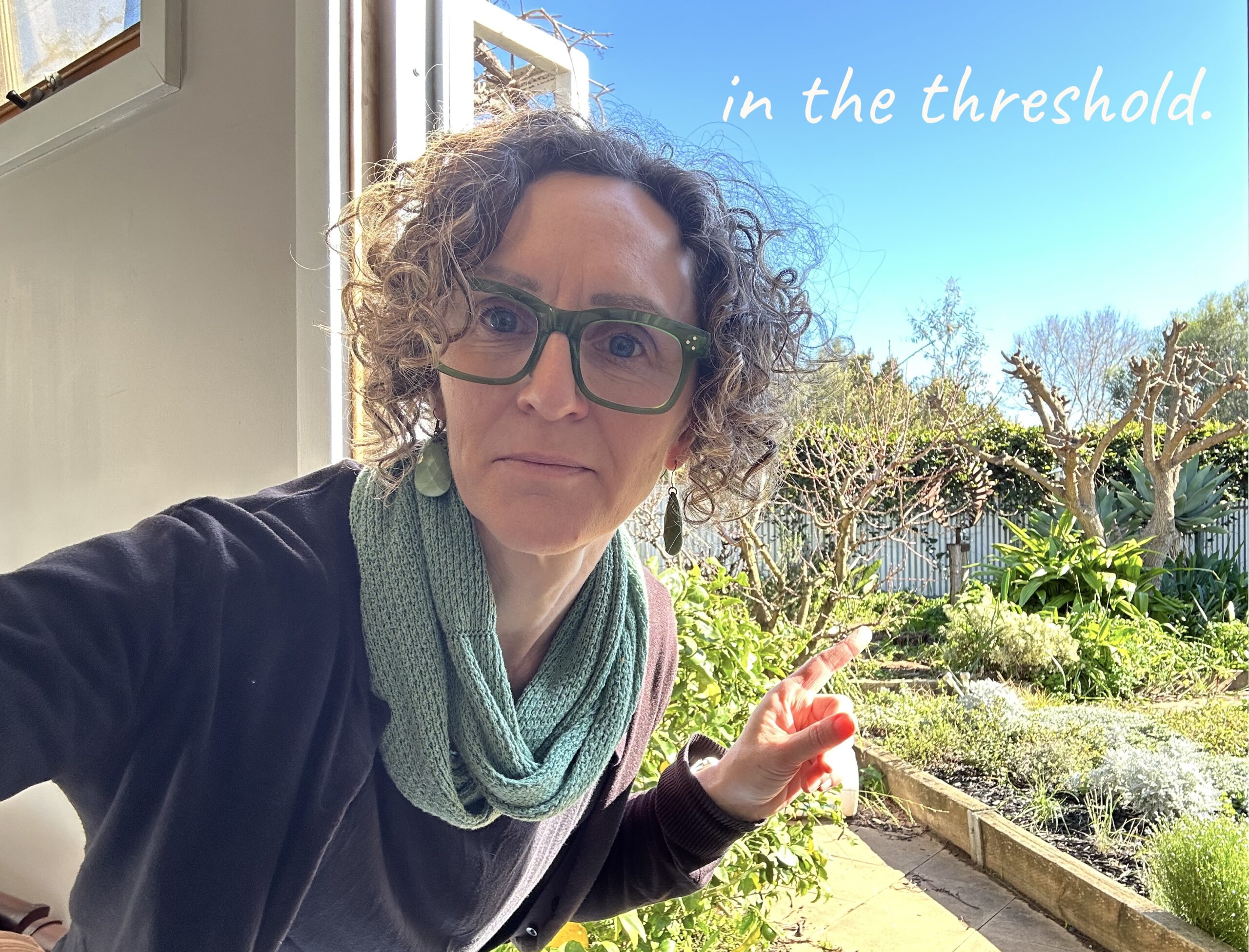 a head and shoulders shot of a woman with short curly brown hair and green glasses leans into a doorway pointing out to a garden behind her. The words 'in the threshold' are written in white handwriting font